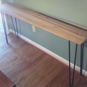 May include: A long, narrow wooden table with a light wood finish and black hairpin legs. The table is sitting on a hardwood floor.