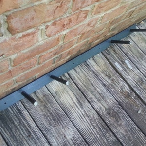 May include: A black metal shelf bracket with four black metal pegs attached to a brick wall. The bracket is mounted to a wooden surface.