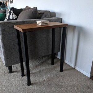 Könnte beinhalten: Kleiner, rechteckiger Beistelltisch mit dunkler Holzplatte und schwarzen Metallbeinen. Der Tisch ist etwa 76 cm hoch und steht neben einem grauen Sofa. Auf der Tischoberfläche liegt ein Stapel quadratischer Untersetzer.