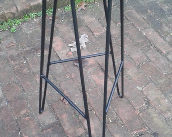 May include: Black metal stool with a round seat and a tripod base. The stool is standing on a brick patio.