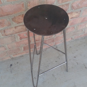 May include: A metal stool with a round, dark grey seat and a triangular base. The stool is made of metal and has a rustic, industrial look.