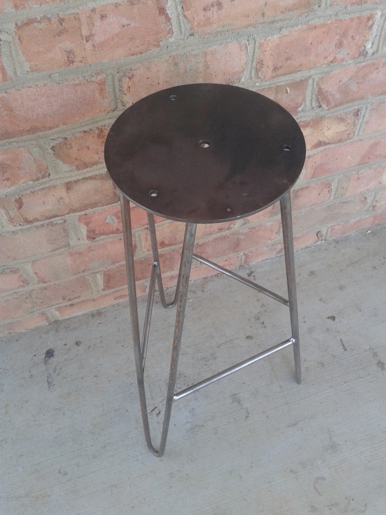 May include: A metal stool with a round, dark grey seat and a triangular base. The stool is made of metal and has a rustic, industrial look.
