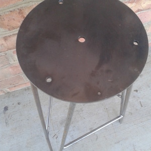 May include: A metal stool with a round, dark grey metal seat and a triangular metal base. The seat has several holes drilled into it.