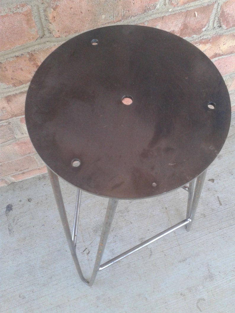 May include: A metal stool with a round, dark grey metal seat and a triangular metal base. The seat has several holes drilled into it.