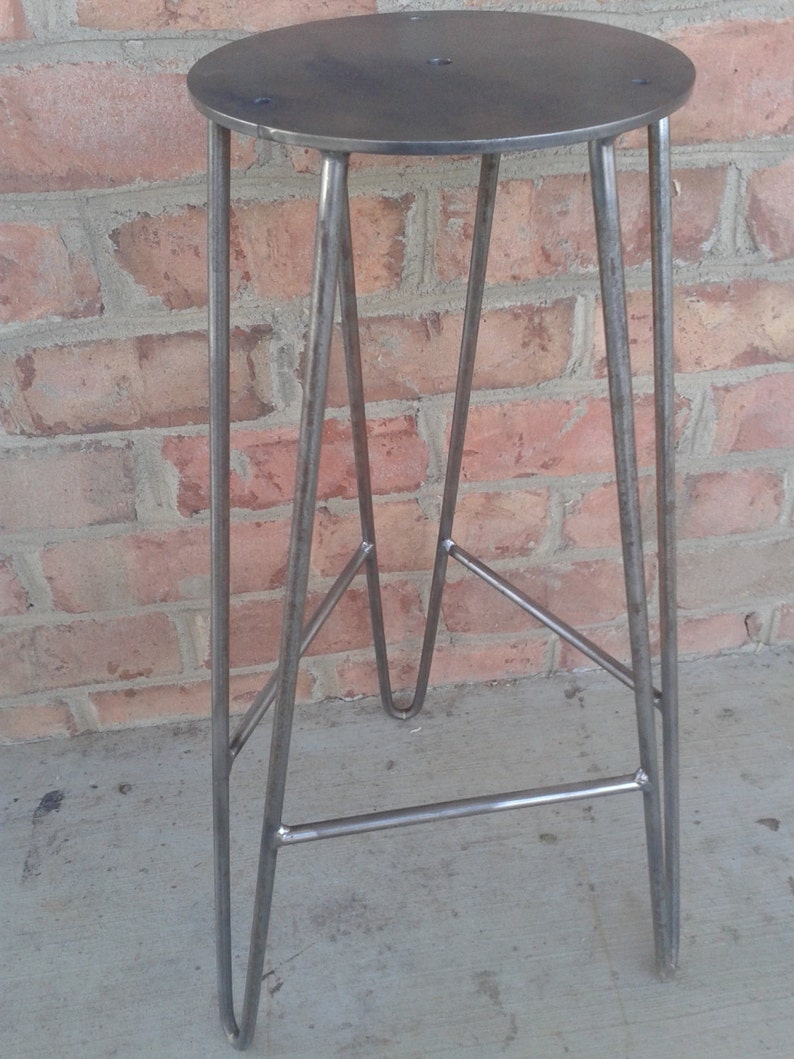 May include: A metal stool with a round, flat top and a simple, industrial design. The stool has four legs that are connected by a crossbar.