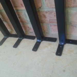 May include: Four black metal shelf brackets with a simple L-shaped design. The brackets are attached to a brick wall and have a flat base.