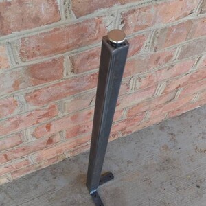 May include: A dark gray metal post with a silver cap, set against a red brick wall. The post has a square profile and is mounted on a concrete surface. The base has two small metal feet.