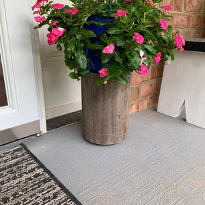 May include: A hanging planter with pink flowers sits on a wooden stand. The planter is blue and the stand is made of wood with a rustic finish.