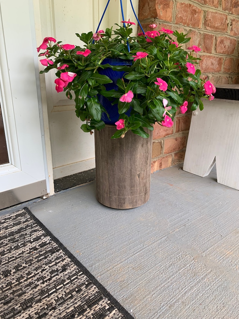 May include: A hanging planter with pink flowers sits on a wooden stand. The planter is blue and the stand is made of wood with a rustic finish.