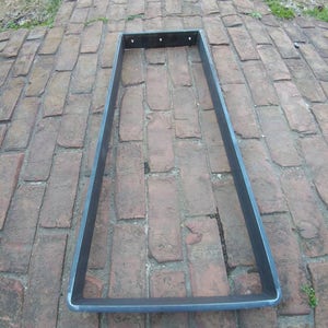 May include: A rectangular metal frame with a dark gray finish. The frame is made of thick metal and has a slightly rounded edge. It is sitting on a brick surface.
