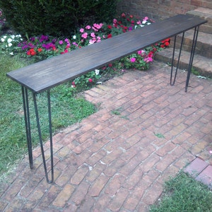 May include: A dark brown wooden console table with black hairpin legs. The table is sitting on a brick patio with a flower garden in the background.