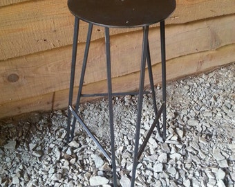 May include: A black metal stool with a round seat and a triangular base. The stool is sitting on a bed of gravel.