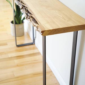 May include: A long, narrow console table with a live edge wood top and black metal legs. The table is made of natural wood with a rustic finish. The legs are simple and modern, creating a contrast with the natural wood top.