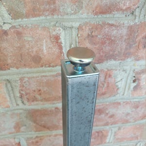 May include: Close-up of a silver metal table leg with a round, silver cap on top. The leg is square and has a textured surface.