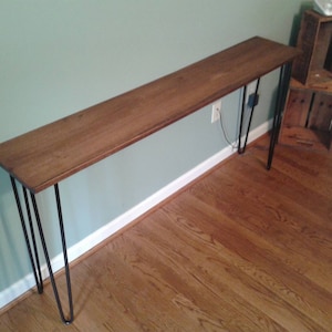 May include: A long, wooden console table with a natural wood finish and black hairpin legs. The table is sitting on a hardwood floor.