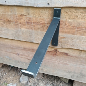 May include: Black metal shelf bracket mounted to a wooden wall. The bracket is angled and has a flat surface for a shelf.