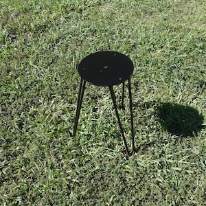 May include: A black metal stool with a round top and three legs. The stool is sitting on a green grassy surface.