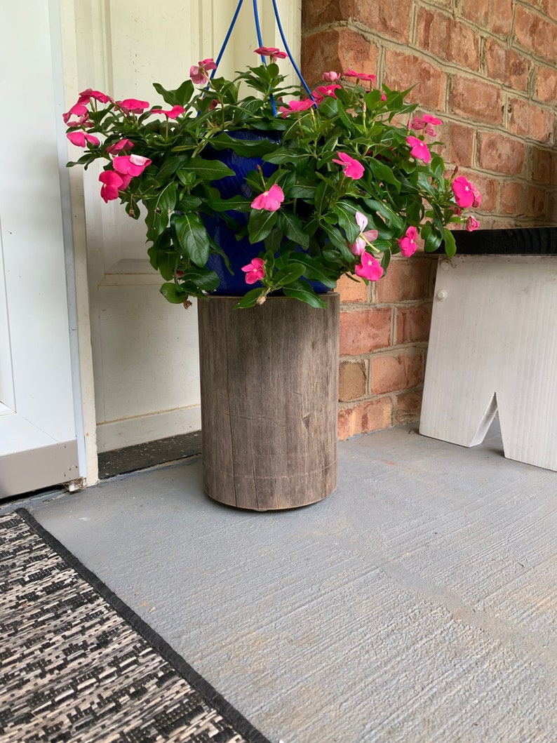 May include: A hanging planter with pink flowers sits on a wooden stand. The stand is made of a dark brown wood and has a rough texture. The planter is blue and has a white rope hanging from it.