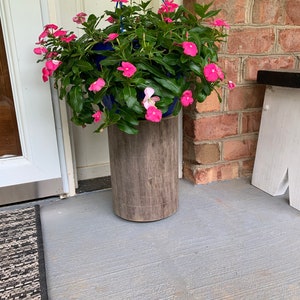 May include: A hanging planter with pink flowers sits on a wooden stand. The planter is blue and the stand is brown. The planter is hanging from a blue rope.
