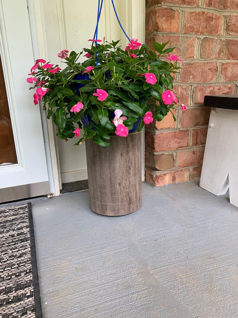 May include: A hanging planter with pink flowers sits on a wooden stand. The planter is blue and the stand is brown. The planter is hanging from a blue rope.