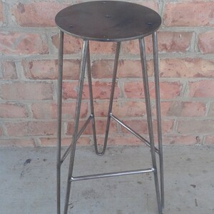 May include: A metal stool with a round, flat top and a simple, industrial design. The stool has three legs that are bent into a hairpin shape.