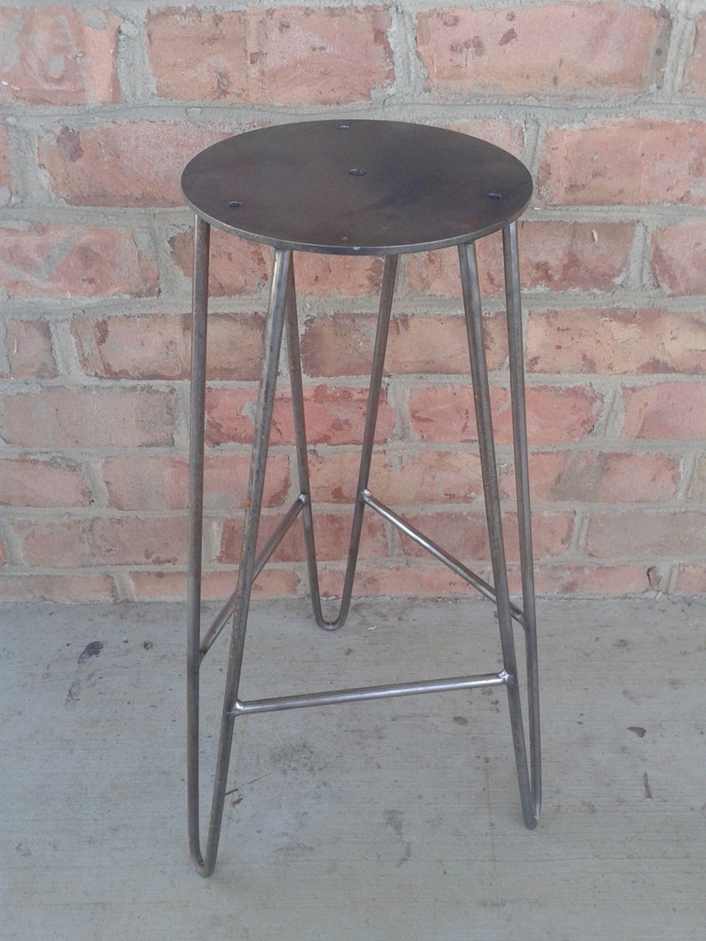 May include: A metal stool with a round, flat top and a simple, industrial design. The stool has three legs that are bent into a hairpin shape.