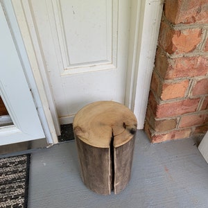 May include: A rustic wooden stool made from a tree trunk with a natural wood finish. The stool has a crack down the center and is sitting on a gray concrete floor.