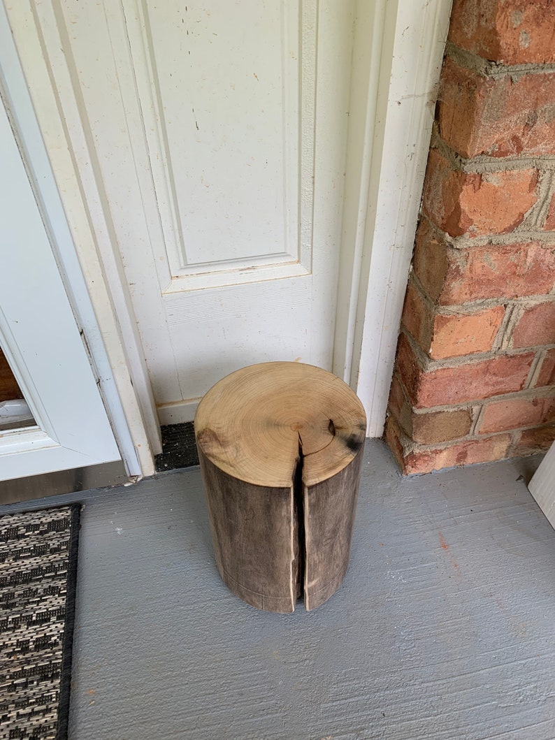 May include: A rustic wooden stool made from a tree trunk with a natural wood finish. The stool has a crack down the center and is sitting on a gray concrete floor.