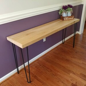 May include: A light wood console table with black hairpin legs. The table is against a purple wall and has a small wooden box with the word "Blessed" on it.