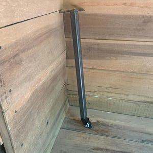 May include: A single, dark gray metal table leg with a small black caster wheel at the bottom. The leg is attached to a wooden structure with a metal plate. The wood has a natural, light brown grain.