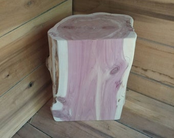 May include: A natural wood stool with a pink and white wood grain pattern. The stool is made from a single piece of wood and has a rustic, organic look.