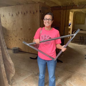 May include: A woman wearing a pink t-shirt with a paw print logo holds a triangular metal bracket. The bracket is made of steel and has a silver finish.