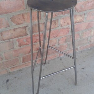May include: A metal stool with a round seat and a triangular base. The stool is made of steel and has a raw, industrial look.