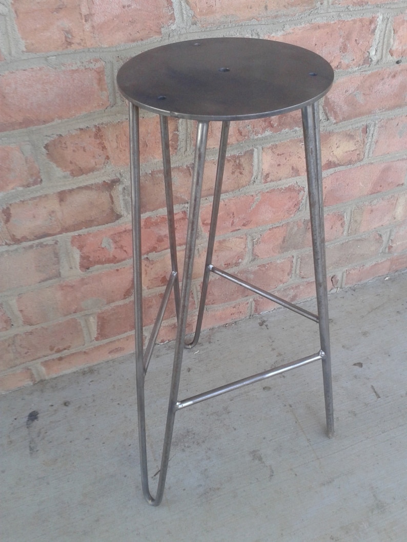 May include: A metal stool with a round seat and a triangular base. The stool is made of steel and has a raw, industrial look.