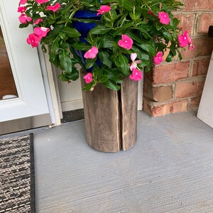 May include: A hanging planter with pink flowers and green leaves sits on a wooden stand. The stand is made of two pieces of wood that are joined together.