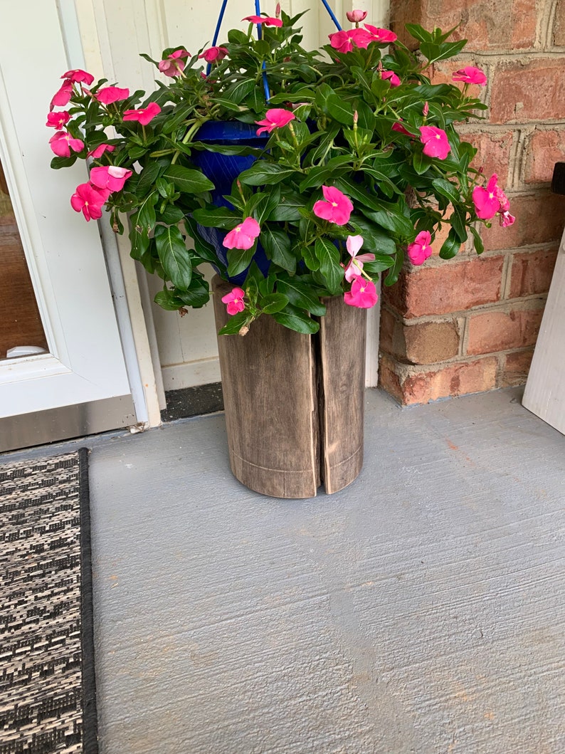 May include: A hanging planter with pink flowers and green leaves sits on a wooden stand. The stand is made of two pieces of wood that are joined together.