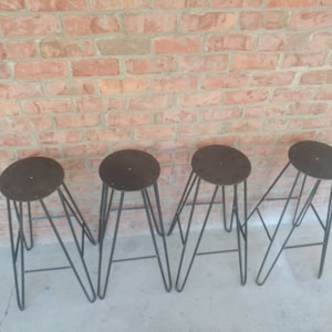 May include: Four black metal bar stools with round brown seats. The stools are arranged in a row on a concrete floor in front of a brick wall.