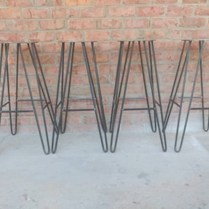 May include: Four black metal hairpin leg stools with a simple design. The stools are arranged in a row on a concrete floor.