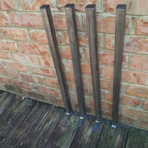 Könnte beinhalten: Vier Metalltischbeine mit quadratischen Sockeln und schwarzer Oberfläche. Die Beine sind an einer Backsteinwand befestigt.