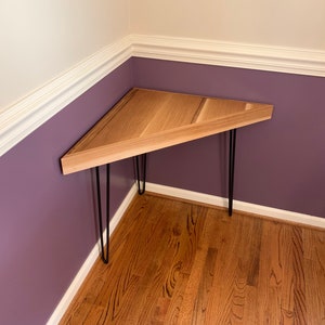 May include: A triangular wooden table with a light wood finish and black hairpin legs. The table is in a corner of a room with purple walls and white trim.