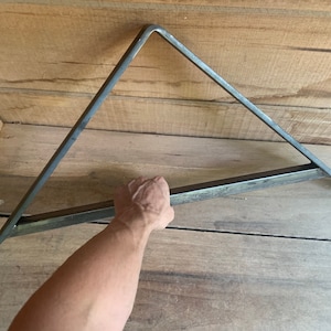 May include: A metal triangle shelf bracket with a flat, rectangular bar across the bottom. The bracket is made of steel and has a raw, unfinished look.