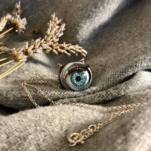 May include: A silver necklace with a round pendant featuring a blue glass eye with a black pupil and eyelashes. The pendant is surrounded by a silver frame.