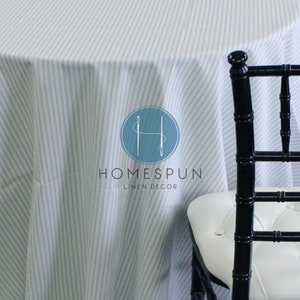 May include: White tablecloth with thin blue stripes. The tablecloth is draped over a round table. A black chair with a white cushion is in the background. The Homespun Linen Decor logo is in the center of the tablecloth.