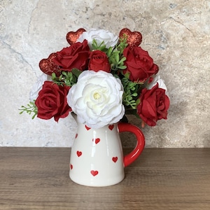 Può includere: Un'anfora di ceramica bianca con cuori rossi stampati, riempita di un bouquet di rose rosse e fiori bianchi. Il bouquet è decorato con cuori rossi glitterati.