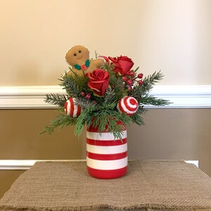 Op de afbeelding: Een kerst-bloemstuk in een rood-wit gestreepte pot. Het arrangement bevat rode rozen, groene dennentakken, rode bessen en een gingerbread man-versiering.