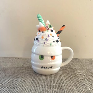 May include: White ceramic mug shaped like a mummy with a green and orange eye, topped with whipped cream, sprinkles, candy corn, and a candy eye. The mug has a stitched mouth and a black and orange striped straw.