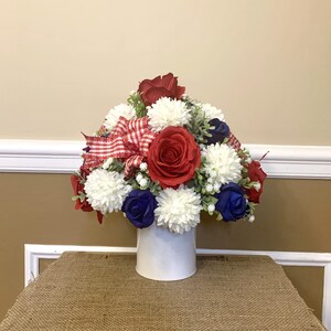 以下が含まれることがあります： 赤、白、青のフラワーアレンジメントが詰まった白い陶器の花瓶。アレンジメントには赤いバラ、白いカーネーション、青いバラ、赤と白のチェックのリボンが含まれています。