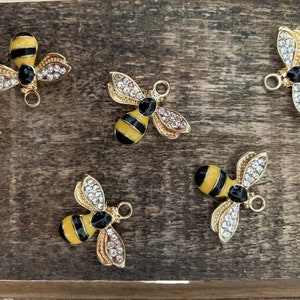 Pode incluir: Cinco pingentes de abelha dourados com listras pretas e amarelas e strass transparentes. As abelhas têm um laço dourado para prender em joias ou outras manualidades.