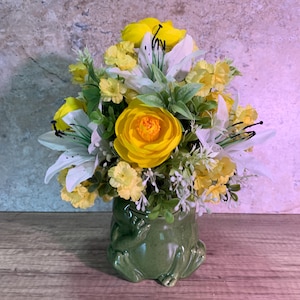 Pode incluir: Um arranjo floral com ranúnculos amarelo-brilhantes e lírios brancos com folhagem verde, colocado num vaso de cerâmica verde-oliva brilhante em forma de sapo com membros visíveis e corpo arredondado, criando uma peça central decorativa.