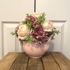 Könnte beinhalten: Eine rosafarbene Teekanne mit einem Strauß weißer und lila Blumen. Die Blumen sind in der Teekanne angeordnet und fallen über die Ränder. Die Teekanne steht auf einem Holztisch.